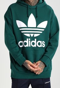 Grön huva i mjukt tyg, med en stor vit Adidas-logga och det karakteristiska utförandet med tre ränder på framsidan. Justerbar huva.