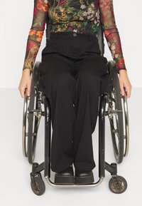 Rullestolbruker med en blomstret langermet topp og svarte vide bukser, hender på håndtakene til rullestolen, iført svarte sko.