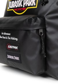 Mochila negra de material sintético suave, con los logotipos de Jurassic Park y Eastpak, bolsillo frontal con cremallera y cordón decorativo.