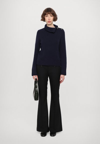 Pull en tricot bleu marine foncé avec un col en entonnoir et des détails de boutons, associé à un pantalon noir évasé. Le mannequin tient un petit sac à main noir.