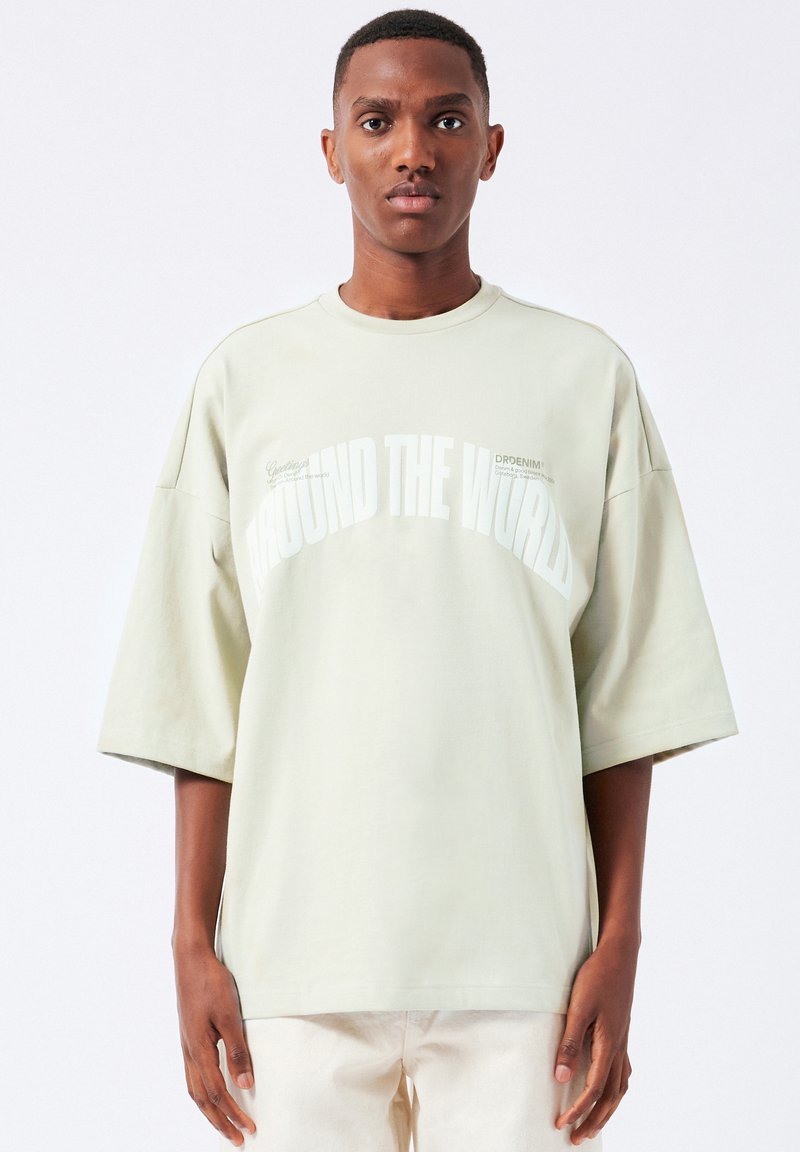 Lys grøn oversized T-shirt lavet af blødt stof, med store hvide bogstaver "AROUND THE WORLD" og små grafikker på forsiden.