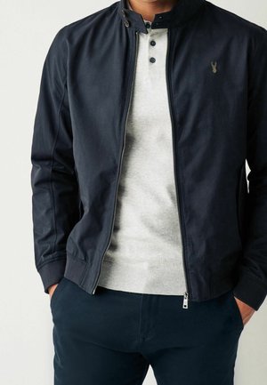 Mężczyzna w granatowej kurtce na zamek błyskawiczny, z jasnoszarym swetrem henley i granatowymi spodniami, z rękami w kieszeniach.