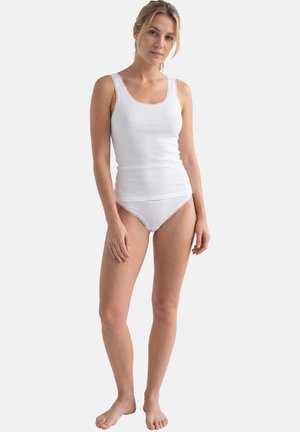 Frau steht barfuß, trägt ein weißes Spitzentanktop und passende Unterwäsche vor einfachem hellem Hintergrund.