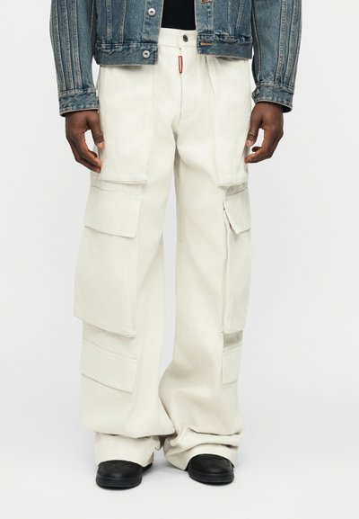 Personne portant un pantalon cargo blanc ample, des chaussures noires et une veste en jean bleue sur un fond clair uni.