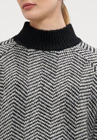 Žena v černobílém svetr s chevronovým vzorem, silným černým žebrovaným stojáčkem, viditelné rty, vlasy stažené dozadu.