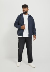 Chaqueta con capucha navy y cierre de cremallera, llevada sobre una camiseta blanca, combinada con pantalones negros y zapatillas blancas. Tejido suave, corte relajado.