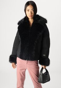 Doudoune noire avec un grand col et des poignets en fausse fourrure, arborant un patch logo sur la manche. Associée à un pantalon rose clair et un sac à main noir.