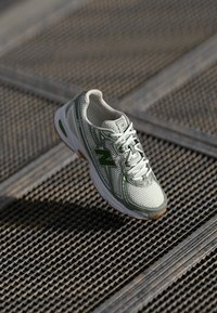 Baskets New Balance blanches et vertes avec tige en mesh, semelle en caoutchouc texturée, système de laçage et logo sur le côté extérieur, posées sur une surface grillagée.