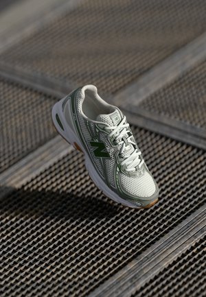 Baskets New Balance blanches et vertes avec tige en mesh, semelle en caoutchouc texturée, système de laçage et logo sur le côté extérieur, posées sur une surface grillagée.