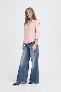 Blusa rosa chiaro con colletto e maniche tre quarti, abbinata a jeans in denim a gamba larga. Tacchi neri a punta completano l'outfit.