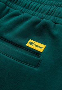 Pantalones de chándal verdes con cintura elástica, que presentan una pequeña etiqueta amarilla "LEVEL UP" y un detalle de bolsillo plano en la parte delantera. Textura suave, diseño casual.