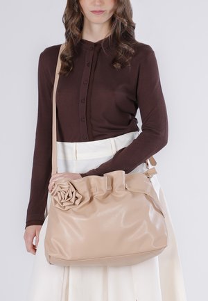 Kvinde iført mørkebrun knappet overdel og hvid nederdel, holder beige skuldertaske med blomsterdetalje i taljehøjde.