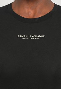Close-up van een persoon die een zwart T-shirt met ronde hals draagt, met witte tekst "ARMANI EXCHANGE MILANO / NEW YORK" op de borst.