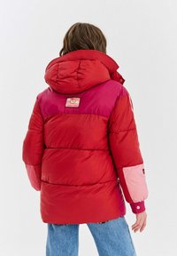 Osoba oblečená v červené a růžové zateplené zimní bundě s kapucí a nášivkou "Sweet Things" na zádech, doplněná modrými džíny.