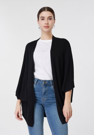 Γυναίκα με σκούρα μαλλιά που φοράει ασημένια σκουλαρίκια κρίκους, λευκό μπλουζάκι, μαύρο χαλαρό ζακέτα και μπλε τζιν ψηλόμεσο, μπροστά σε απλό φόντο.