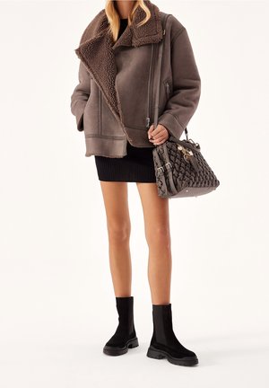 Vrouw gekleed in een bruine schapenvachtjas, zwarte minirok, zwarte enkellaarsjes, en een getextureerde donkergrijze handtas dragend, op een witte achtergrond.