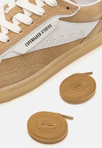Barna suéd sneaker fehér bőr részletekkel és perforált mintával. Fehér fűzőkkel rendelkezik, és tartalmaz egy pár azonos barna fűzőt is. Átlátszó talp.