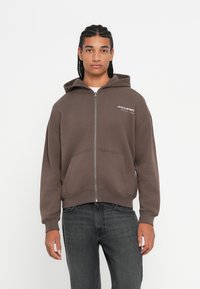 Mladý muž s vrkôčkami, ktorý má na sebe hnedú zipsovú mikinu McLaren a tmavosivé džínsy, stojí pred obyčajným bielym pozadím.