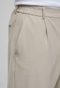Pantalons beiges avec une taille élastique, dotés de poches latérales et d'un tissu lisse et léger. La conception plissée apporte de la structure.