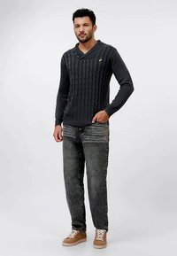 Tmavosivý sveter s pleteným vzorom a šálovým golierom, skombinovaný so vyblednutými čiernymi džínami a svetlohnedými teniskami s bielymi šnúrkami. Jednoduchý žltý detail loga.