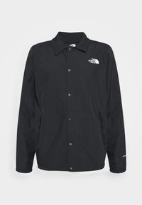 Veste coupe-vent noire à bouton avec col, poignets élastiques, poches latérales et logo blanc The North Face sur la poitrine et la manche.
