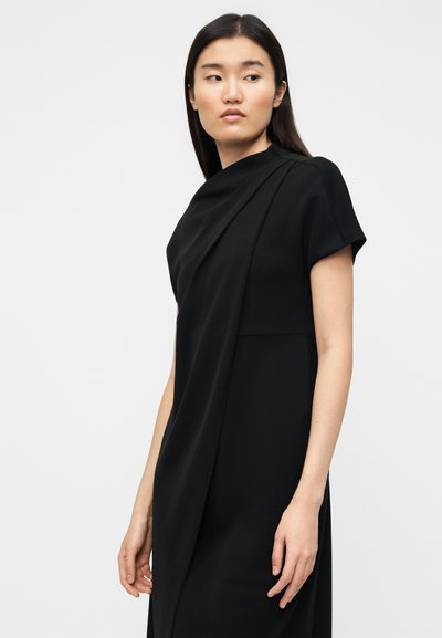 Μαύρο φόρεμα με κοντό μανίκι τύπου καπελού, με σχεδιασμό μπροστινής ζώνης, απαλό ύφασμα και μήκος έως το γόνατο. Απλή σιλουέτα χωρίς ορατά σχέδια ή διακοσμητικά στοιχεία.