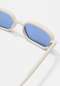 Zonnebril met een crème-kleurig kunststof frame, blauwe lenzen en gestructureerde pootjes met reliëfdruk van het merk "GUESS". Rond model met een slank design.