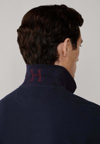 Marinblå pikétröja med hög krage och en vinröd "H"-logotyp på baksidan. Materialet verkar vara texturerat och kragen är vikt.