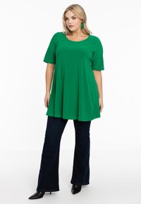 Groene tuniek met korte mouwen en een ronde halslijn, losvallend tot de heupen, gecombineerd met donkere flared jeans en puntige zwarte enkellaarzen.