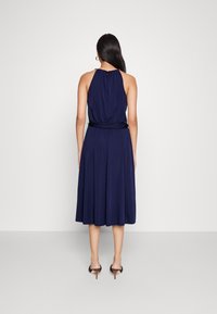 Kvinna med långt mörkt hår som bär en ärmlös midiklänning i marinblått och svarta klacksandaler, står mot en enfärgad ljus bakgrund.