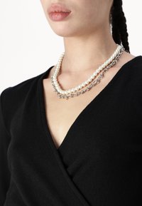 Driedubbele ketting met witte parels en zilverkleurige accenten, gedragen over een zwart V-hals top, die het halsgebied accentueert.