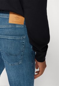 Jeans de denim azul con corte recto, que presentan un parche de marca de cuero marrón, costuras visibles y contornos de los bolsillos frontales. Camisa negra de manga larga.