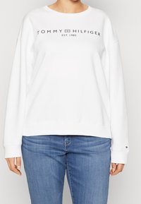 Sudadera blanca con cuello redondo, que presenta un logo negro de Tommy Hilfiger y el año de fundación impreso en el pecho. Textura suave.
