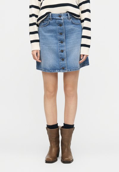 Femme portant une jupe en denim bleu boutonnée sur le devant, un pull rayé, des chaussettes noires et des bottes en cuir marron mi-mollet, debout devant un fond blanc.