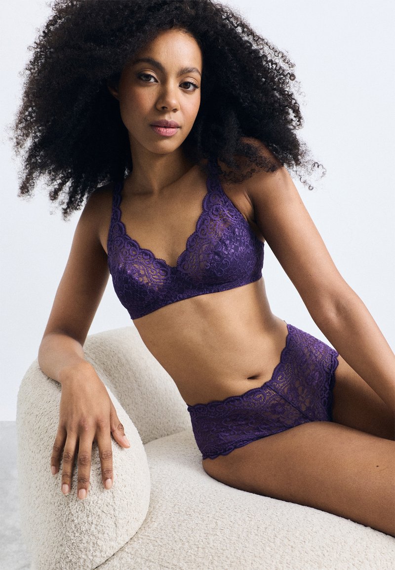 Set di bralette e slip abbinati in pizzo viola con bordi a ondina, caratterizzato da intricati motivi floreali. La modella posa seduta su una sedia beige testurizzata.