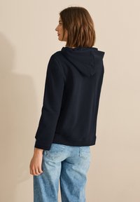 Sudadera con capucha de color navy, de manga larga, con corte relajado y puños acanalados, combinada con jeans de tiro alto de color azul claro. Textura de tejido suave.
