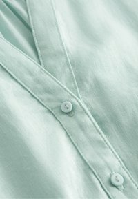 Light mint green linen shirt featuring a V-neckline, visible stitching, and a front button with a matching buttonhole.