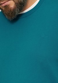 Türkisfarbener Strickpullover mit Rundhalsausschnitt und gerippten Details, versehen mit einem dezenten Logo auf der Brust. Der Stoff wirkt glatt und körpernah.