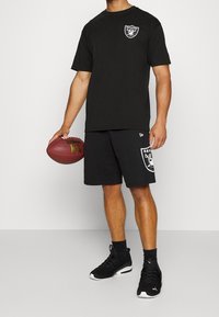 Svart bomullströja med Raiders-logotyp på vänster bröst, tillsammans med svarta shorts med en Raiders-grafik. Svarta träningsskor och en fotboll hålls.