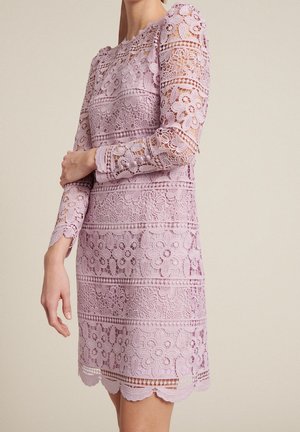 Liliowa sukienka koronkowa z kwiatowymi wzorami, długimi rękawami i falbankowanym dołem, noszona przez osobę na jasnym, jednolitym tle.