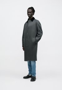 Grå lang frakke med en sort krave, der har knapper foran og sidelommer, kombineret med blå jeans og sorte sko. Glat tekstur.