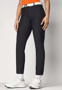 Fekete golf nadrág, slim fit szabású, rugalmas anyagból készült. Előre zsebekkel rendelkezik, cipzáras bokája van, és egy fehér övvel van stílusosan párosítva.