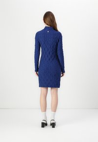 Rochie tricotată albastră cu mâneci lungi, având un model texturat și un guler înalt. Are un mic detaliu cu nasture la spate.