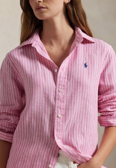 Kvinde iført en lyserød og hvid skjorte med lodrette striber, knappet op foran, med op rullede ærmer og en lille blå broderet logo på brystet.