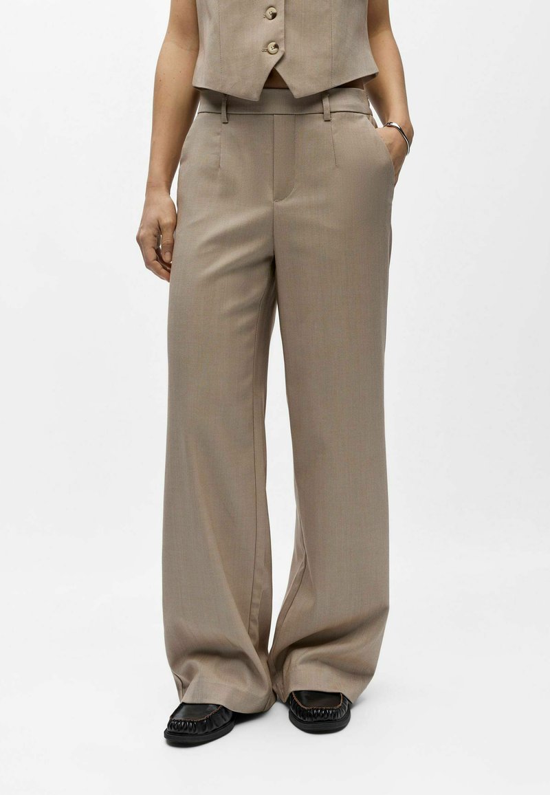 Femme portant un pantalon beige à jambes larges et taille haute avec poches latérales, assorti à un gilet boutonné de la même couleur et des mocassins noirs.