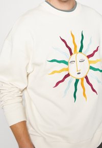 Cremefarbener Pullover mit einem in Gelb, Grün, Rot und Blau gestickten Sonnendesign, das ein Gesicht und wellenförmige Strahlen zeigt.