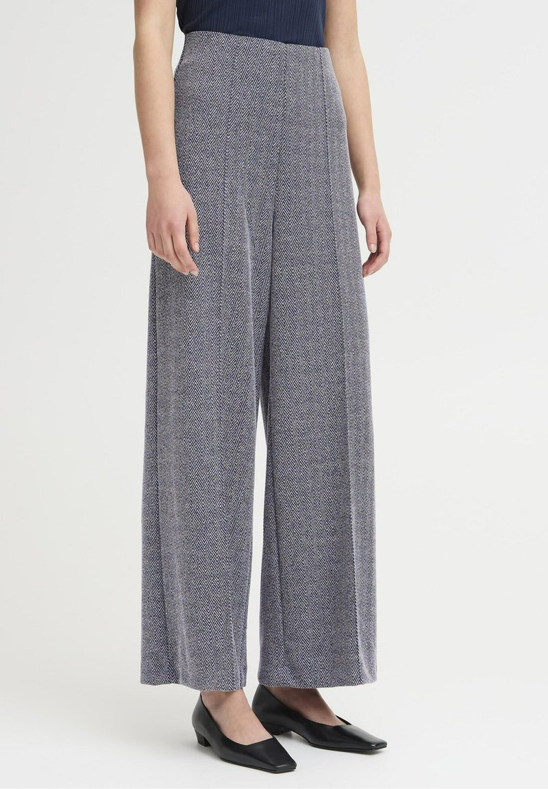 Femme portant un pantalon taille haute à jambes larges avec motif chevrons bleu et blanc, associé à des chaussures noires à talons bas.