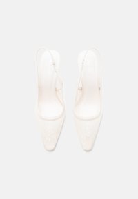 Ζευγάρι μπεζ slingback τακούνια με μυτερές μύτες και διακριτικό σχέδιο από λουλουδάτη δαντέλα, εμφανιζόμενα από πάνω σε λευκό φόντο.