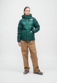 Ženska v zeleni puhovki North Face, ohlapnih rjavih hlačah in sivih športnih copatih, stoji z eno roko v žepu na enobarvni beli ozadju.
