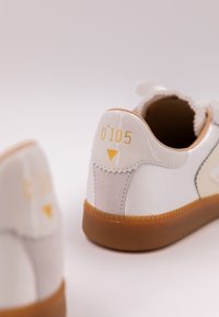 Vita sneakers med beige mockadetaljer, gummisula och "O'105"-logotyp i gult på hälen. Slät, blank yta.